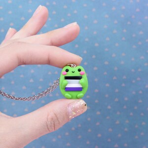 Puede incluir: Un collar con un dije de rana verde que sostiene una bandera del orgullo asexual p&uacute;rpura, blanca y negra. La rana sonr&iacute;e y tiene mejillas rosadas.