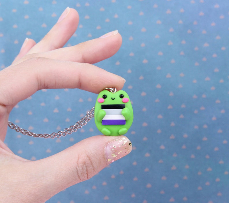 Puede incluir: Un collar con un dije de rana verde que sostiene una bandera del orgullo asexual p&uacute;rpura, blanca y negra. La rana sonr&iacute;e y tiene mejillas rosadas.