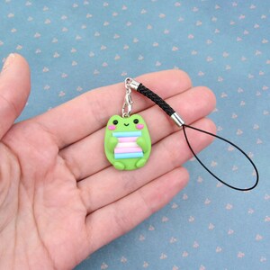K&ouml;nnte beinhalten: Gr&uuml;ner Frosch-Anh&auml;nger mit einem Transgender-Pride-Flaggen-Design, befestigt an einem schwarzen Handyband.