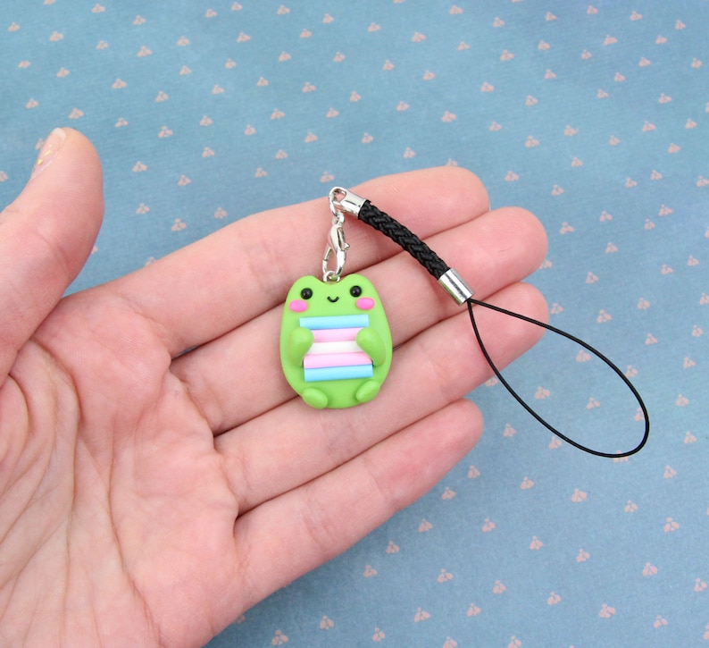 K&ouml;nnte beinhalten: Gr&uuml;ner Frosch-Anh&auml;nger mit einem Transgender-Pride-Flaggen-Design, befestigt an einem schwarzen Handyband.