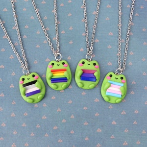 K&ouml;nnte beinhalten: Vier silberne Kettenhalsb&auml;nder mit gr&uuml;nen Frosch-Anh&auml;ngern, die verschiedene Pride-Flaggen halten. Die Flaggen sind Regenbogen, lila, schwarz und wei&szlig; sowie hellblau, rosa und wei&szlig;.