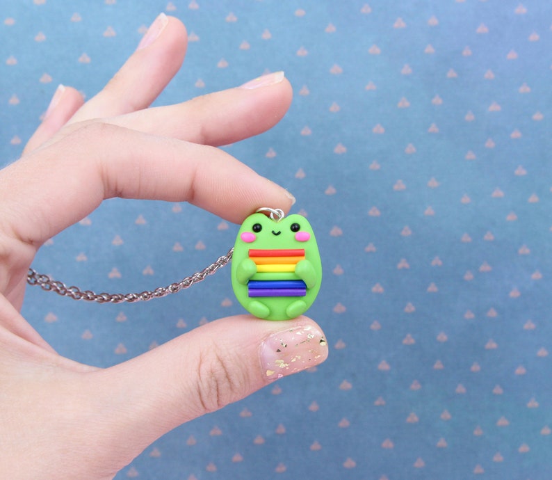 Puede incluir: Un collar con un dije de rana verde que sostiene una bandera arco&iacute;ris del orgullo.