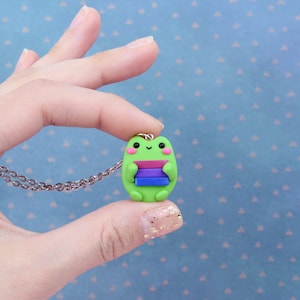 Puede incluir: Un collar con un dije de rana verde que sostiene una pila de libros con rayas de color rosa, morado, azul y blanco. La rana tiene una peque&ntilde;a sonrisa y mejillas rosadas.