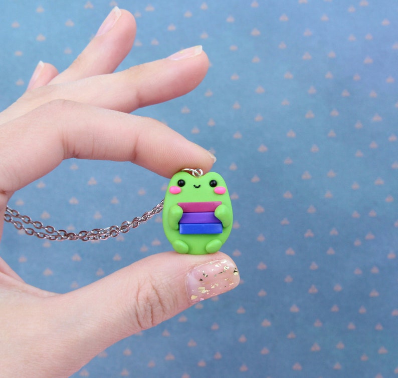 Puede incluir: Un collar con un dije de rana verde que sostiene una pila de libros con rayas de color rosa, morado, azul y blanco. La rana tiene una peque&ntilde;a sonrisa y mejillas rosadas.