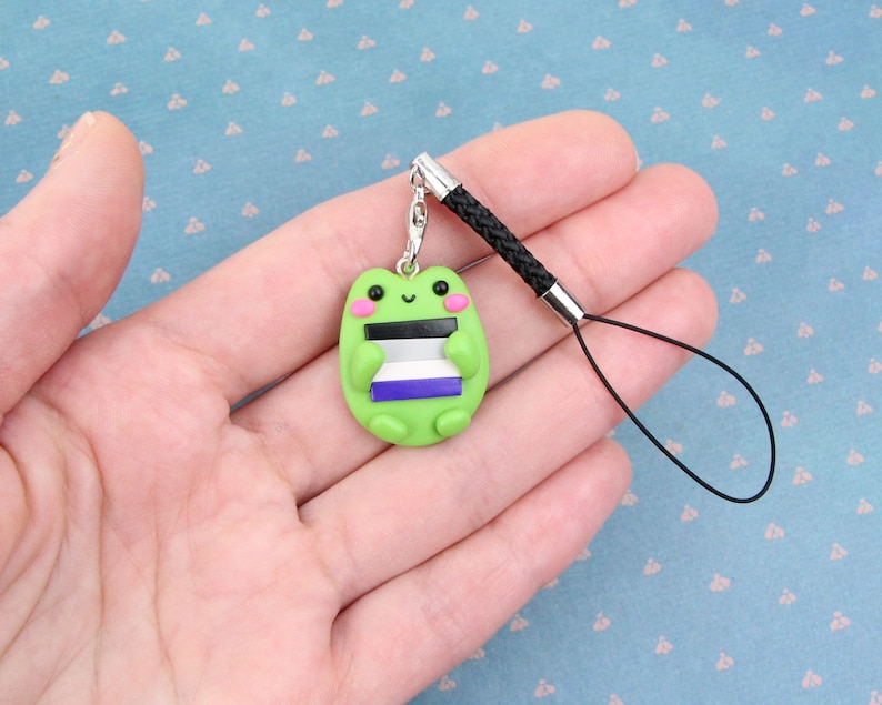 K&ouml;nnte beinhalten: Ein gr&uuml;ner Frosch-Anh&auml;nger mit rosafarbenem Gesicht und schwarzen Augen, der eine kleine Flagge mit schwarzen, wei&szlig;en, lila und grauen Streifen h&auml;lt. Der Anh&auml;nger ist an einem schwarzen Handyband befestigt.