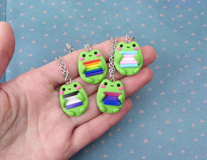 Puede incluir: Cuatro collares de rana verde con diferentes colores de banderas del arco&iacute;ris. Cada rana sostiene una pila de rayas de colores que representan a la comunidad LGBTQ+.