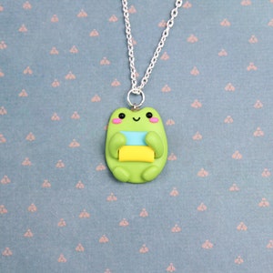 May include: A green frog necklace with a smiling face and holding a stack of colorful blocks. The necklace is on a blue background with a repeating pattern.