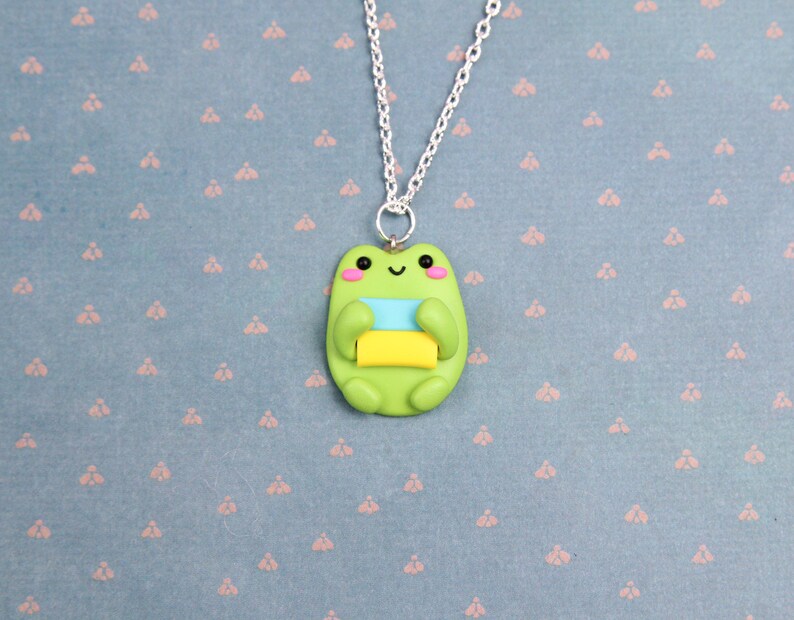 May include: A green frog necklace with a smiling face and holding a stack of colorful blocks. The necklace is on a blue background with a repeating pattern.