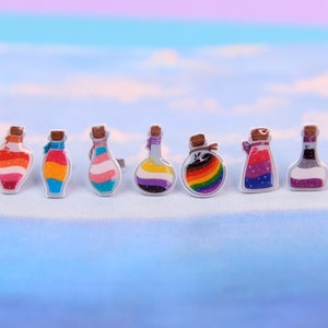 May include: Seven small glass bottles with cork stoppers and colourful liquid inside. Each bottle features a different pride flag design, including lesbian, bisexual, transgender, pansexual, genderfluid, and asexual.