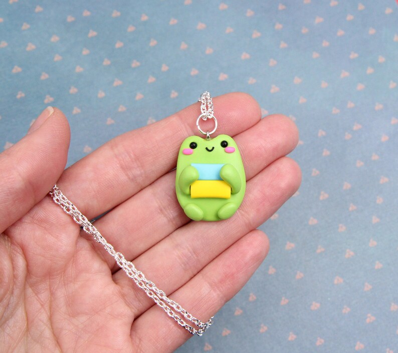 May include: A silver chain necklace with a green frog charm holding a stack of colorful paper. The frog has a happy expression with black eyes and a pink blush.