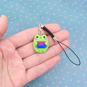 K&ouml;nnte beinhalten: Gr&uuml;ner Frosch-Anh&auml;nger mit einer Bi-Pride-Flagge an einem schwarzen Handyband.