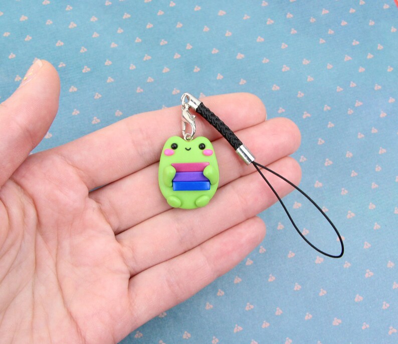 K&ouml;nnte beinhalten: Gr&uuml;ner Frosch-Anh&auml;nger mit einer Bi-Pride-Flagge an einem schwarzen Handyband.