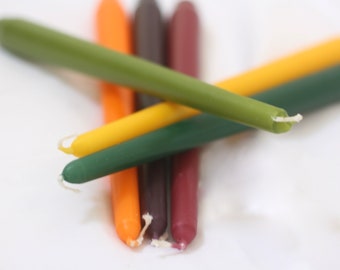 May include: A group of five taper candles in various colors, including green, yellow, orange, and burgundy. The candles are arranged in a criss-cross pattern.