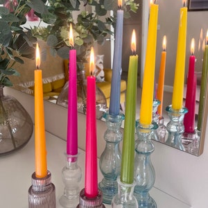 May include: A collection of six lit taper candles in vibrant colours including orange, magenta, pink, grey, lime green, and yellow, each standing in individual clear or tinted glass candle holders with ribbed or smooth pedestal designs.
