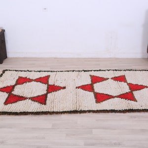 K&ouml;nnte beinhalten: Ein wei&szlig;er Teppich mit einem roten und braunen geometrischen Muster aus Sternen. Der Teppich hat einen Fransenrand.