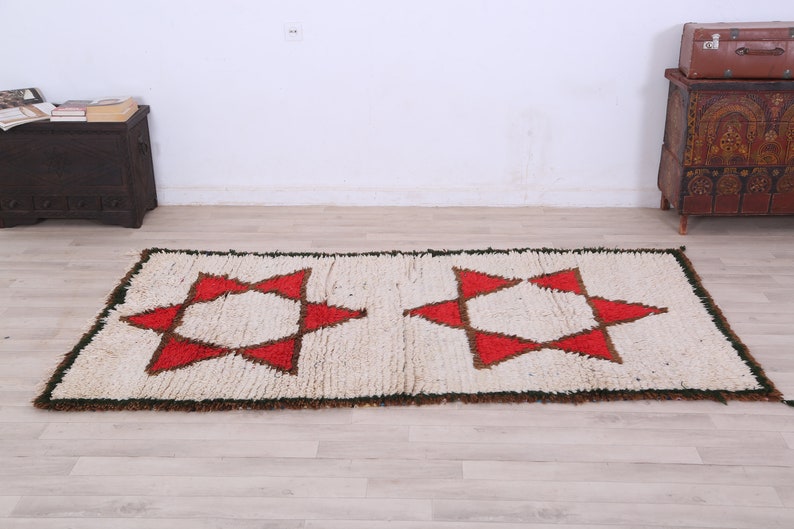 K&ouml;nnte beinhalten: Ein wei&szlig;er Teppich mit einem roten und braunen geometrischen Muster aus Sternen. Der Teppich hat einen Fransenrand.