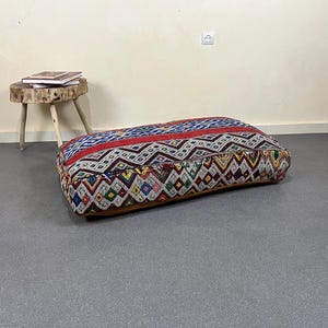 May include: A rectangular floor cushion with a colorful geometric pattern. The cushion features a red stripe and a brown leather base. A small wooden stool with books on top is in the background.