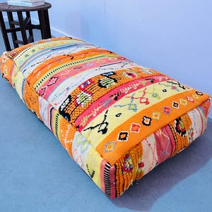 May include: A colorful, patterned pouf with orange, yellow, pink, and blue hues. The pouf is made of woven fabric and has a square shape.