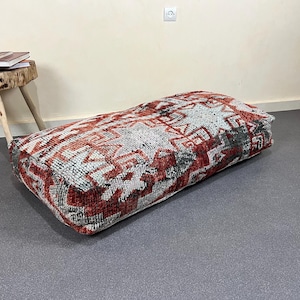 May include: Rectangular floor cushion with a geometric pattern in red, white, and black. The cushion is placed on a gray surface. A small wooden side table with books on top is in the background.