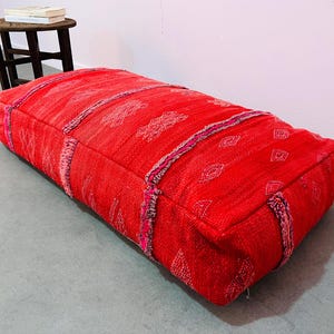 May include: A rectangular, red floor cushion with geometric patterns and decorative trim. The cushion is made of woven fabric and has a soft, inviting appearance. The cushion is approximately 36 inches long, 24 inches wide, and 8 inches high.