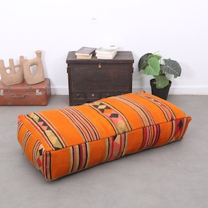 May include: An orange rectangular floor cushion with a striped pattern in red, black, and yellow. The cushion has a geometric design on the ends. Other items in the background include a brown trunk, decorative objects, and a potted plant.