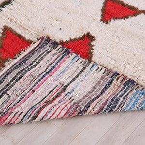 K&ouml;nnte beinhalten: Ein wei&szlig;er, flauschiger Teppich mit roten und braunen geometrischen Mustern. Die Unterseite des Teppichs ist mit bunten Streifen gewebt.