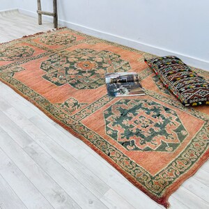 May include: A vintage rug with a traditional Persian design in shades of pink, green, and brown. The rug has a floral pattern and is laid out on a light wood floor. A decorative pillow with a geometric pattern is placed on the rug.