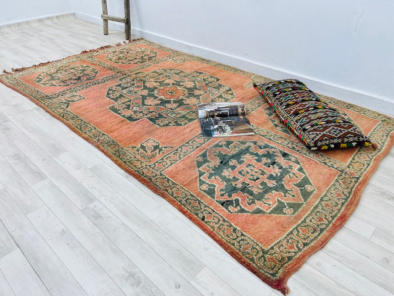 May include: A vintage rug with a traditional Persian design in shades of pink, green, and brown. The rug has a floral pattern and is laid out on a light wood floor. A decorative pillow with a geometric pattern is placed on the rug.