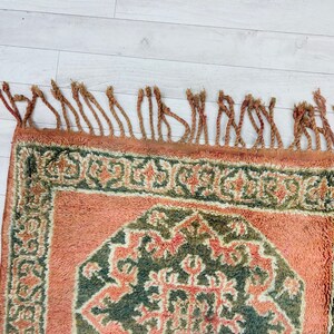 May include: A vintage rug with a geometric pattern in shades of green, brown, and orange. The rug has a fringe border.