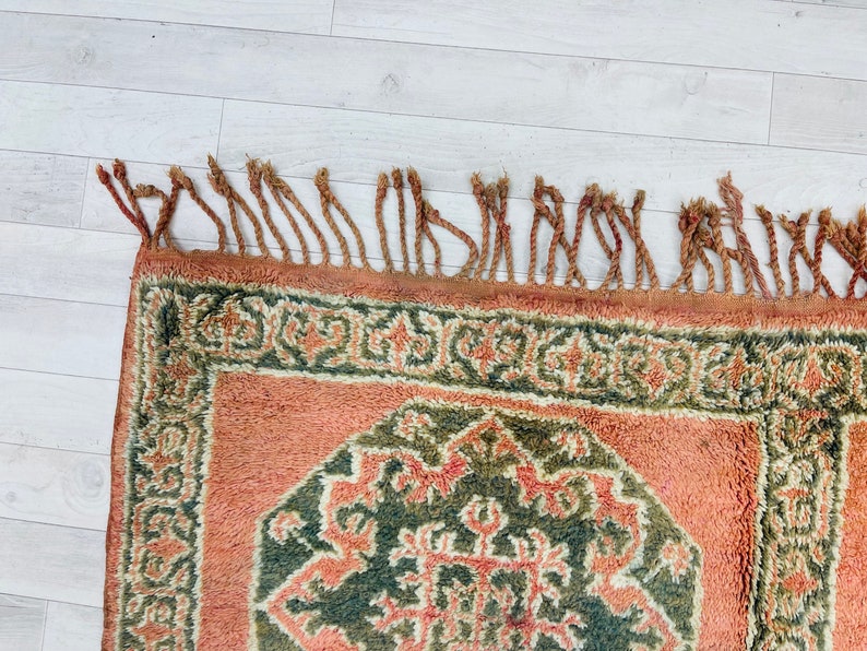 May include: A vintage rug with a geometric pattern in shades of green, brown, and orange. The rug has a fringe border.