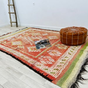 Puede incluir: Una alfombra con estampado rojo, blanco y verde con un puf de cuero marr&oacute;n en la parte superior. La alfombra tiene un borde de flecos y est&aacute; hecha de un material tejido.