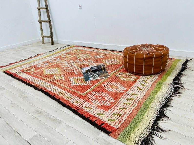 Puede incluir: Una alfombra con estampado rojo, blanco y verde con un puf de cuero marr&oacute;n en la parte superior. La alfombra tiene un borde de flecos y est&aacute; hecha de un material tejido.