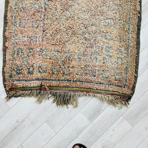 May include: A vintage rug with a faded, cream-colored background and a geometric pattern in shades of blue, red, and brown. The rug has a fringe border and is slightly worn.