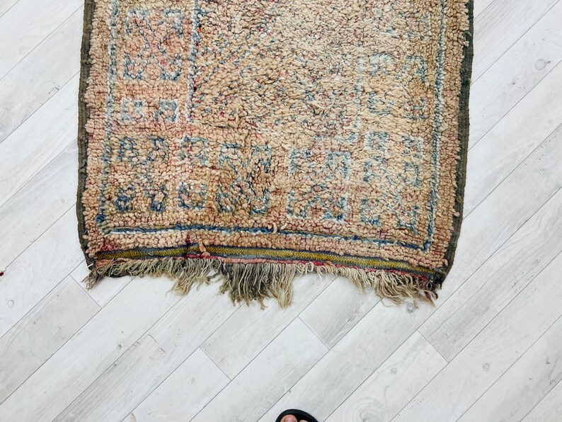 May include: A vintage rug with a faded, cream-colored background and a geometric pattern in shades of blue, red, and brown. The rug has a fringe border and is slightly worn.