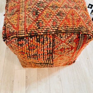 K&ouml;nnte beinhalten: Ein quadratischer, rot-orange-schwarzer Pouf-Ottomane. Der Pouf ist aus gewebtem Stoff gefertigt und hat eine getuftete Textur.