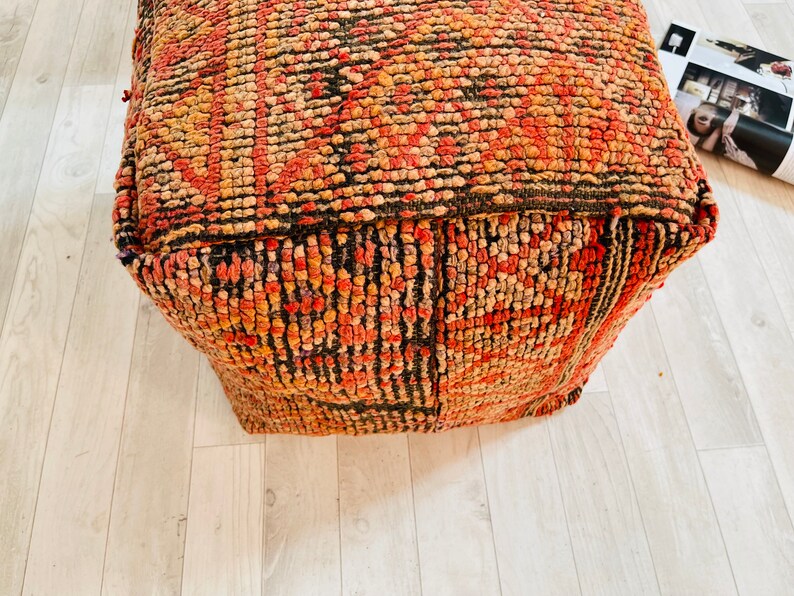 K&ouml;nnte beinhalten: Ein quadratischer, rot-orange-schwarzer Pouf-Ottomane. Der Pouf ist aus gewebtem Stoff gefertigt und hat eine getuftete Textur.
