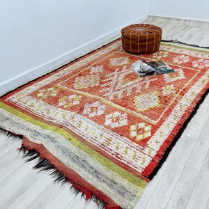 Puede incluir: Una alfombra con estampado rojo, blanco y amarillo con borde de flecos. Un puf de cuero marr&oacute;n se encuentra sobre la alfombra.