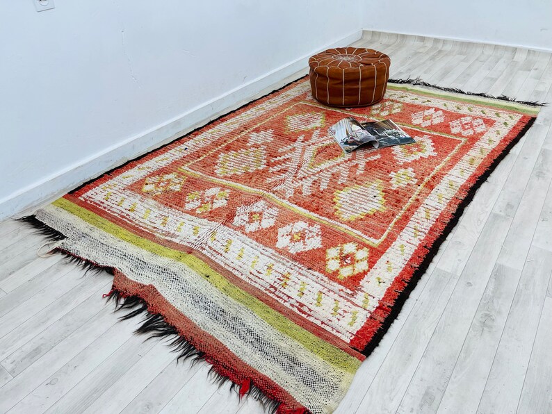 Puede incluir: Una alfombra con estampado rojo, blanco y amarillo con borde de flecos. Un puf de cuero marr&oacute;n se encuentra sobre la alfombra.