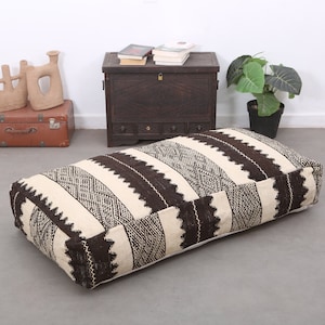 May include: Rectangular floor cushion with a cream and brown striped pattern. The cushion is placed on a gray floor. Other items in the background include a wooden chest, a brown suitcase, and decorative pottery.