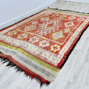 Puede incluir: Una alfombra roja, blanca y negra con un dise&ntilde;o geom&eacute;trico. La alfombra tiene un borde de flecos y est&aacute; hecha de lana.