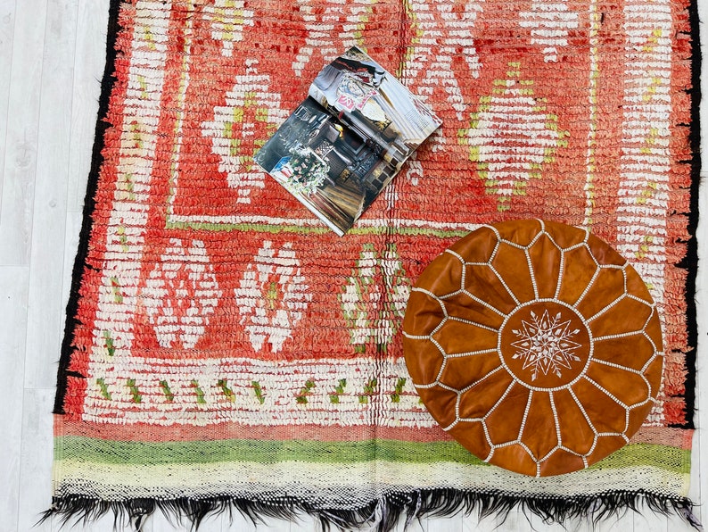 Puede incluir: Una alfombra roja y blanca con un patr&oacute;n con un puf de cuero marr&oacute;n con costuras blancas. Una revista est&aacute; sobre la alfombra.