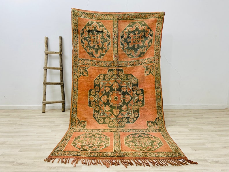May include: A vintage rug with a geometric pattern in shades of orange, green, and brown. The rug has a fringe border and is made of wool.