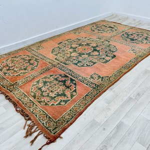 May include: A vintage rug with a peach background and intricate green floral patterns. The rug has fringe on all sides.