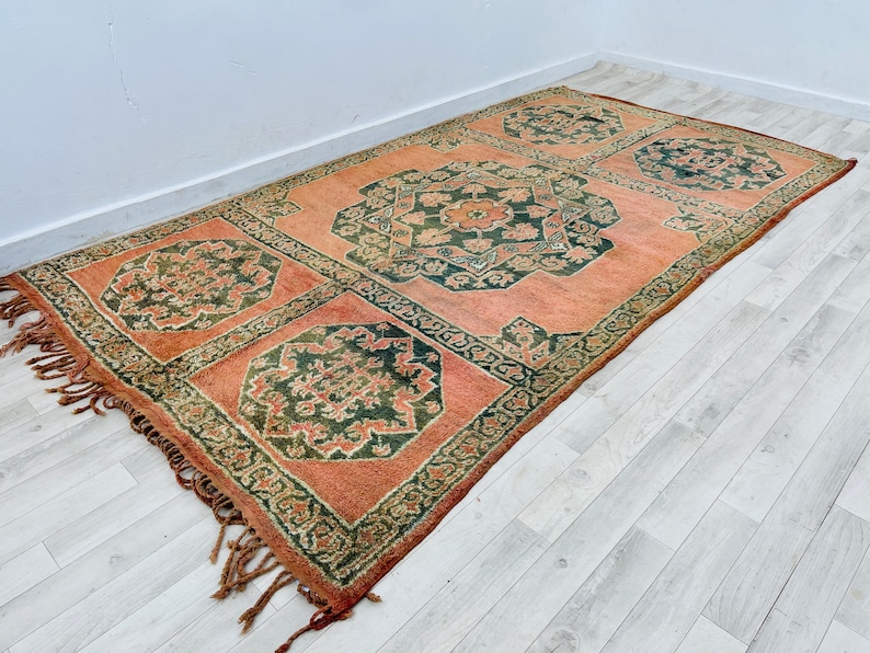 May include: A vintage rug with a peach background and intricate green floral patterns. The rug has fringe on all sides.