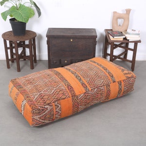 May include: A large, orange and brown patterned floor cushion with a geometric design. The cushion is made of woven fabric and has a square shape.