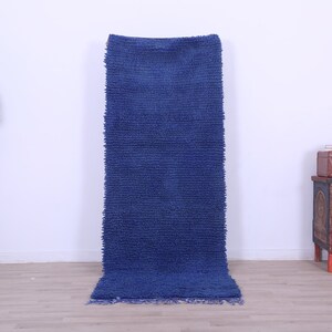 May include: A long, blue, shag rug with a fringe border. The rug is hanging on a white wall.