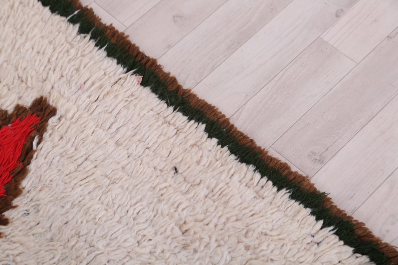 K&ouml;nnte beinhalten: Nahaufnahme eines wei&szlig;en, flauschigen Teppichs mit braunem und gr&uuml;nem Rand. Der Teppich hat ein geometrisches Muster in den Farben Rot, Braun und Wei&szlig;.