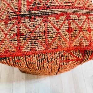 K&ouml;nnte beinhalten: Ein quadratischer Pouf mit rotem, braunem und schwarzem Muster. Der Pouf ist aus einem gewebten Stoff mit einer strukturierten Oberfl&auml;che.