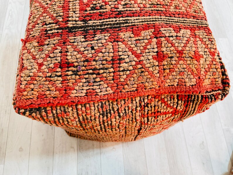 K&ouml;nnte beinhalten: Ein quadratischer Pouf mit rotem, braunem und schwarzem Muster. Der Pouf ist aus einem gewebten Stoff mit einer strukturierten Oberfl&auml;che.