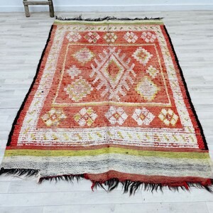 Puede incluir: Una alfombra roja, blanca y verde con un dise&ntilde;o geom&eacute;trico. La alfombra tiene un borde de flecos y est&aacute; hecha de lana.